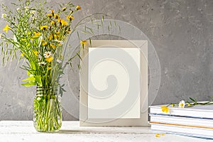 Mock up creation. White frame on white wooden table with grey concrete background, books, notebook and wild flowers