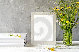 Mock up creation. White frame on white wooden table with grey concrete background, books, notebook and wild flowers