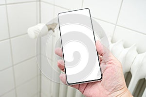 Mock up, hand holding a smartphone with blank white display in front of a heater radiator, concept for rising costs due to energy