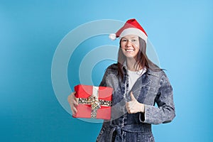 mock up of happy Asian young woman in Santa Claus hat holding a red gift box and giving a thumbs up on a blue background