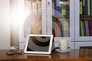 Mock up image of white tablet pc with black blank screen and coffee cup on wooden table in blur office or bookcase background. Re