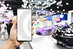 Mock up mobile phone. Hand holding smartphone with  Blurred image of public event exhibition hall showing cars and automobiles.