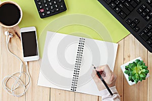 Mock up of office table desk workspace with hands writing on blank notebook and smart phone, calculator, computer keyboard and cof