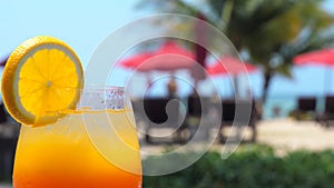 Mocktail drink with orange slice standing in poolside beach bar with sea, palm trees and sun lounges on background