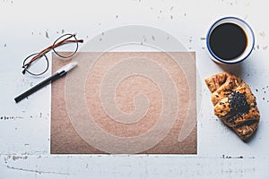 Mockup blank craft sheet of blank craft paper, pen, eye glasses and morning coffee cup with croissant on white wooden