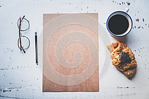 Mockup blank craft sheet of blank craft paper, pen, eye glasses and morning coffee cup with croissant on white wooden