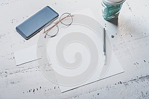 Mockup of blank sheet paper, smartphone, eye glasses and two empty business cards on white wooden desk. Business empty