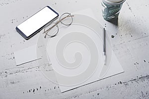 Mockup of blank sheet paper, smartphone, eye glasses and two empty business cards on white wooden desk. Business empty