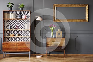 Mockup of empty gold frame above cabinet in grey retro living room interior with lamp. Real photo