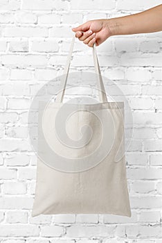 Mockup of female hand holding a blank Tote Canvas Bag on white brick wall background.