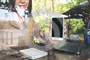 Business woman holding and showing white mobile phone with blank black screen while using laptop