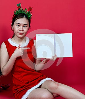 Mockup Image of an Asian Woman Presenting a Book With White Blank Cover Wearing Christmas Attire on a Red Background