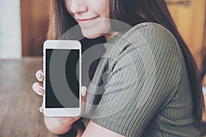 A beautiful woman with smiley face holding and showing white mobile phone with blank black screen in vintage wooden cafe