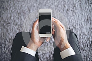 Mockup image of a business man sitting and holding white mobile phone with blank black screen with gray rug