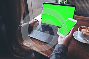 Mockup image of business woman holding mobile phone with blank green screen while using laptop on wooden table