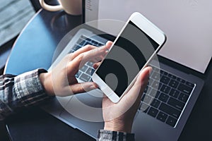 Mockup image of business woman holding white mobile phone with blank black screen with laptop on wooden table