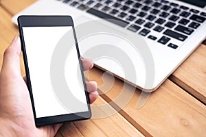 Mockup image of hand holding black mobile phone with blank white screen and silver color laptop on vintage wooden table