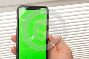 Mockup image of hand holding black mobile phone with green screen on the office table on the background with stationery tools.