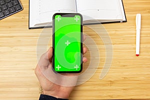 Mockup image of hand holding black mobile phone with green screen on the office table on the background with stationery tools.