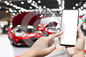 Mockup image of hand holding blank screen mobile and pointing to smart phone with blurred background of new cars display in