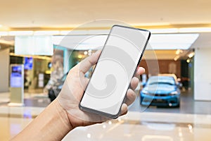 Mockup image of hand holding blank screen mobile smart phone with blurred background of new cars display in showroom, buying new