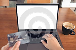 Mockup image of a hand holding credit card while using and typing on laptop with blank white screen and coffee cup