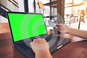 Mockup image of a hand holding credit cards while using and typing on laptop with blank green screen and coffee cup