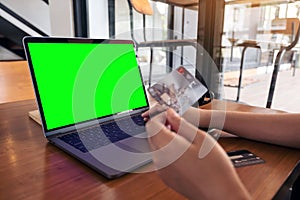 Mockup image of a hand holding credit cards while using and typing on laptop with blank green screen on wooden table