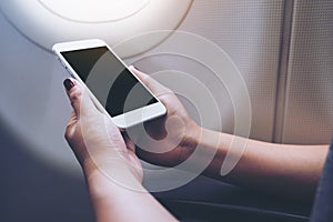 Mockup image of a hand holding and looking at white smart phone with blank desktop screen next to an airplane window