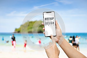 Mockup image of a hand holding and showing modren mobile smart phone  in front of the beach in summer with may tourist and blue