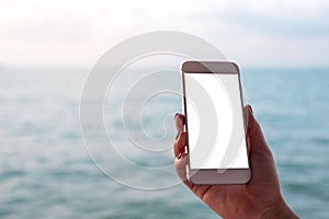 Mockup image of a hand holding and showing white mobile phone with blank black desktop screen in front of the sea and blue sky