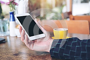 Mockup image of a hand holding white mobile phone with blank black desktop screen and yellow coffee cup on wooden table
