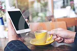 Mockup image of a hand holding white mobile phone with blank black desktop screen and yellow coffee cup on wooden table