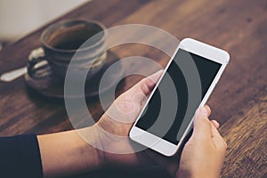 Mockup image of hand holding white mobile phone with blank black screen and coffee cup