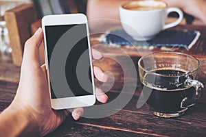 Mockup image of hand holding white mobile phone with blank black screen with coffee cup on wooden table