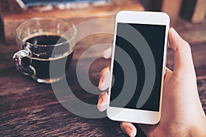 Mockup image of hand holding white mobile phone with blank black screen with coffee cup on wooden table