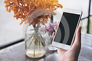 Mockup image of a hand holding white mobile phone with blank black screen with laptop and flower vase on vintage wood table