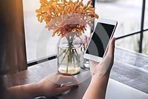 Mockup image of a hand holding white mobile phone with blank black screen with laptop and flower vase on vintage wood table