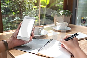 Hand holding white mobile phone with blank desktop screen while writing on notebook with notebook and coffee cup