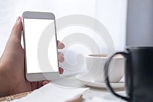 A hand holding white mobile phone with blank screen , black and white coffee cups on vintage wooden table and curtain background