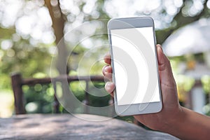 Mockup image of hand holding white mobile phone with blank screen and coffee cup on wooden table