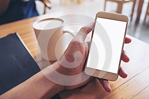 Mockup image of hand holding white mobile phone with blank screen with coffee cup on wooden table