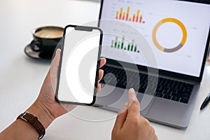 Hands holding a black mobile phone with blank screen while using laptop computer in office