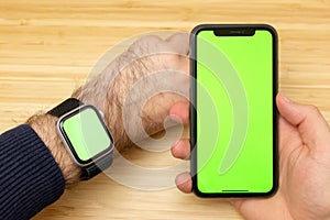 Mockup image of hands holding black mobile phone with green screen and electronic smart watch on wrist . Office table on the.