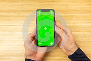 Mockup image of hands holding black mobile phone with green screen on the office table on the background with copy space.