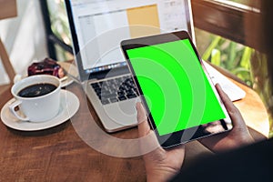 Mockup image of hands holding black tablet pc with blank green screen , laptop , coffee cup and cake on wooden table