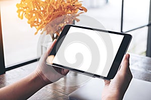 Mockup image of hands holding black tablet pc with blank white screen and flower vase on wooden table