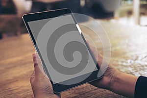 Mockup image of hands holding black tablet pc with gray blank screen on wooden table background