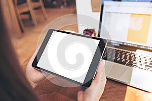 Mockup image of hands holding black tablet pc with white blank screen and laptop on wooden table background