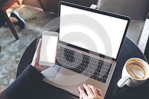 Mockup image of hands holding blank mobile phone while using laptop with blank white desktop screen on table